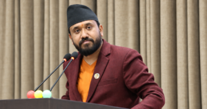 कालो टोपी, मरुन कोट र सुन्तला रङको सर्ट लगाएका सांसद खगेन्द्र सुनार पोडियममा माइक्रोफोनअगाडि उभिएको तस्बिर।
